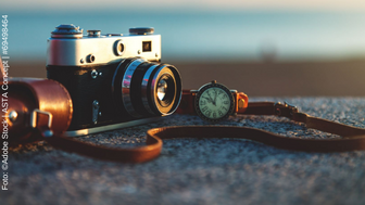Eine Kamera mit Lederband und einer Uhr mit Lederarmband liegen auf einer Steinfläche. Im Hintergrund ist ein Strand zu erkennen.  